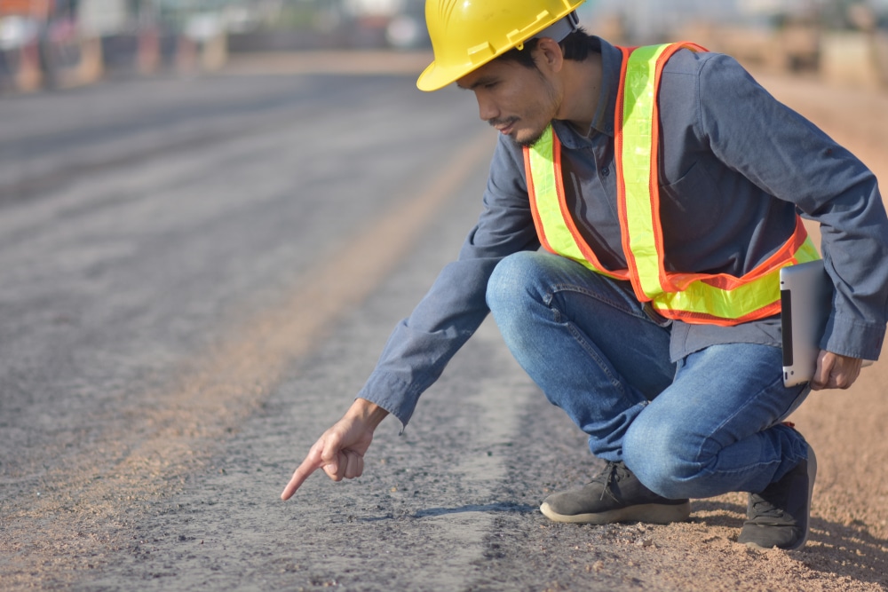 Inspector in a hard hat crouching to examine a cracked asphalt road surface